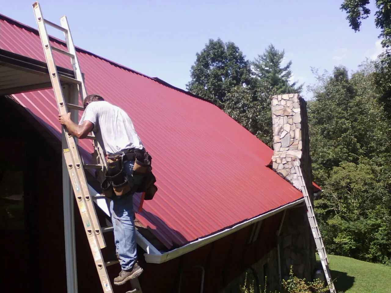 Roofer on a ladder beside a red metal roof and stone chimney — Soffit & Fascia Repair by Roofing Brothers in Hillrose